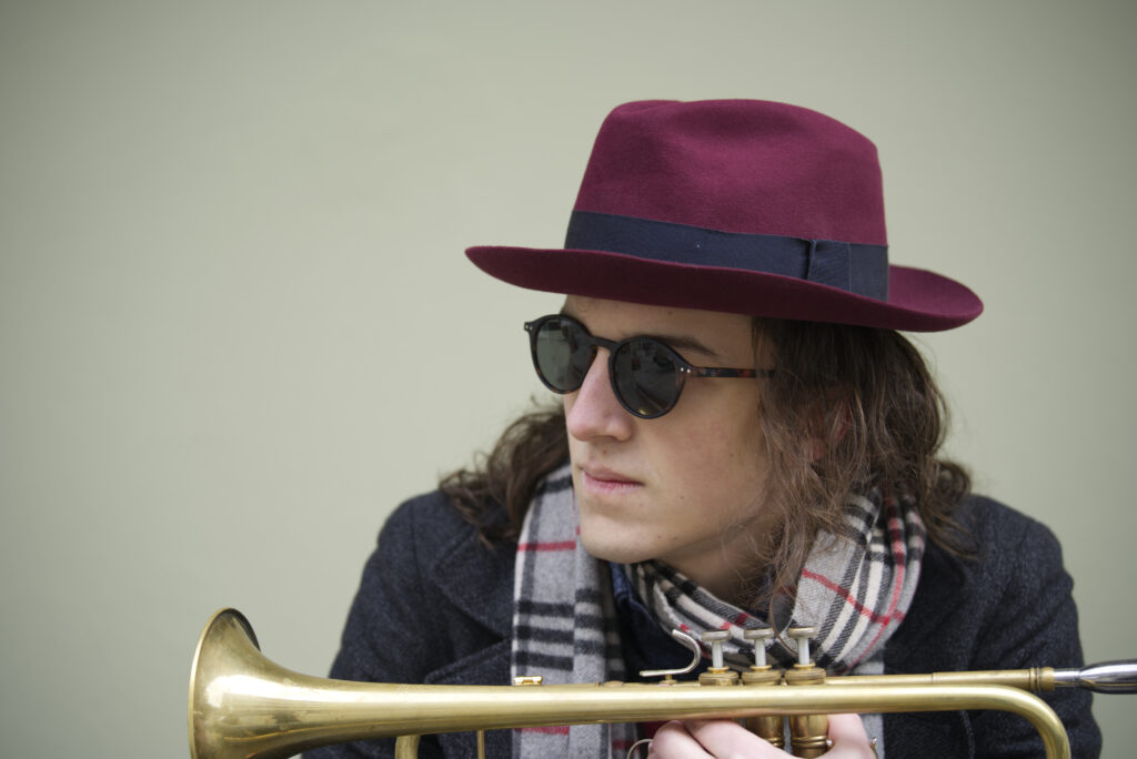 Media-Photo-4 Carlo Bechtel is wearing a fedora-hat and stylish sunglasses. He is looking to the left of the photo whilst holding his instrument in front of him.