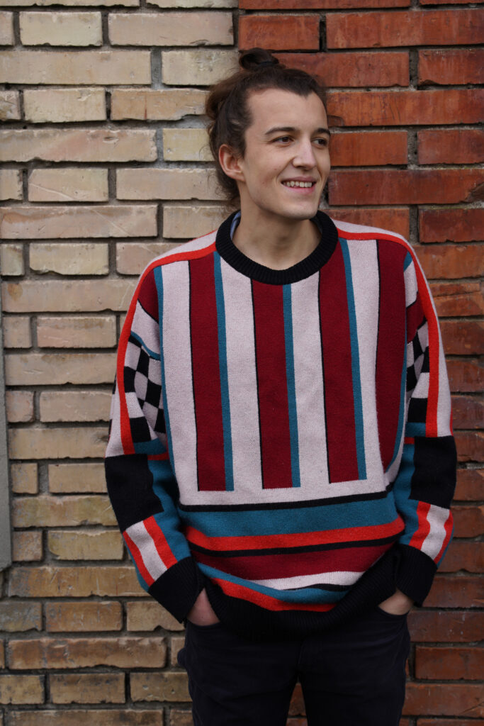 Media-Photo-6 Carlo is standing in front of a two colored brick wall, his hand are in his pockets. He is wearing a colorful sweater and black jeans.