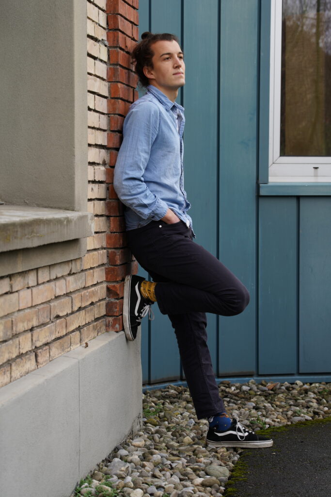 Media-Photo-7 Carlo Bechtel is leaning against a wall with one foot up against the wall. He is looking at something outside the frame.