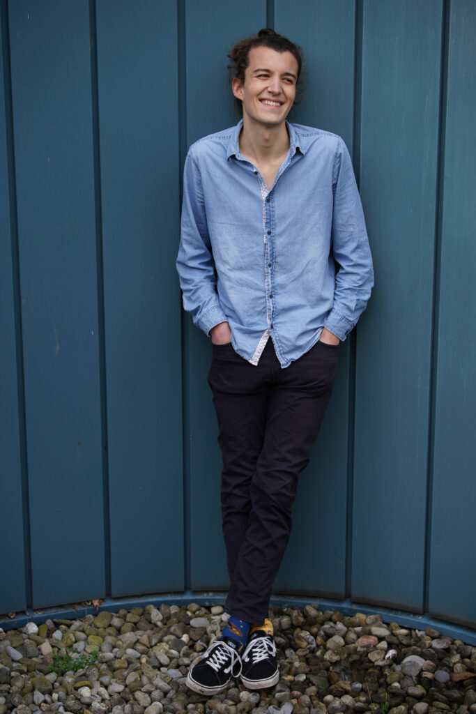 Media-Photo-8 Carlo Bechtel, wearing colorful socks with instruments and music notes on it, is casually leaning against a blue wall.