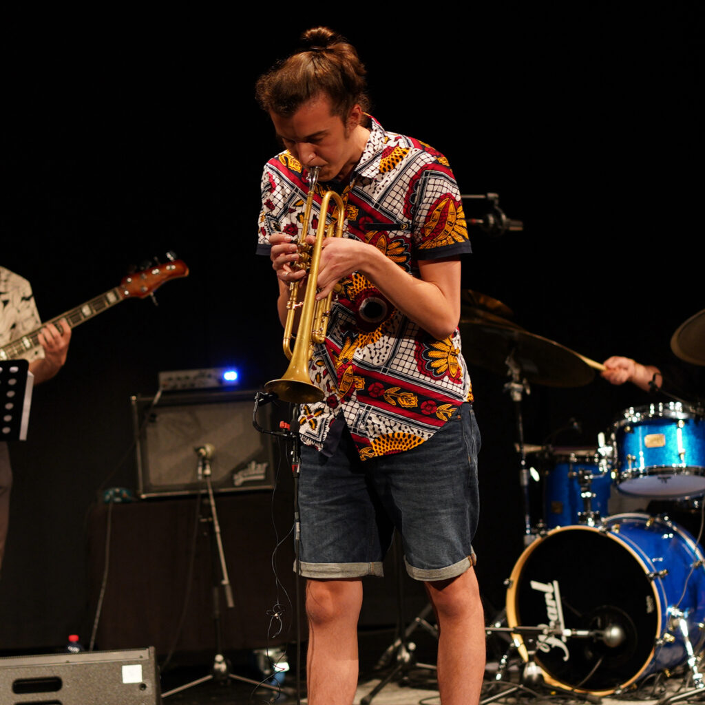 Media-Photo-9 Swiss trumpeter Carlo Bechtel is playing the trumpet at his bachelor-concert in 2021. He is wearing an African shirt. In the background you can see a drum set as well as the neck of an electric bass.